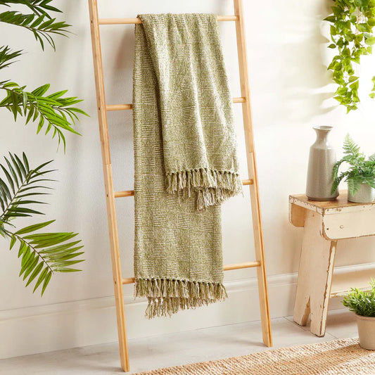 A light green vegan woven throw blanket with fringe hangs on a wooden ladder, surrounded by indoor plants and natural décor in a bright room.