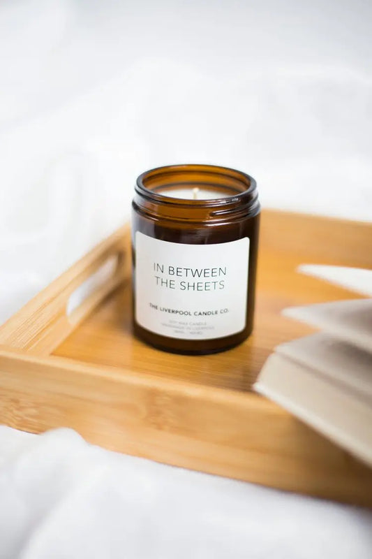 A brown glass soy wax candle labelled In Between The Sheets sits on a wooden tray beside the edge of an open book, filling the air with relaxing notes of lavender, chamomile and vanilla bean.