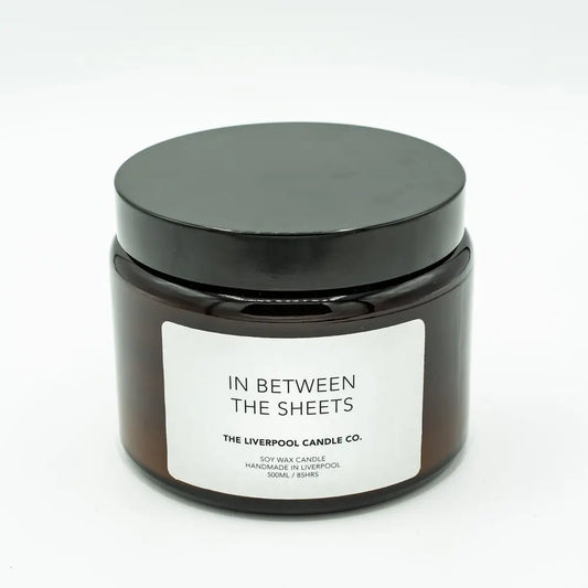 A brown glass jar soy wax candle with a black lid, labelled In Between The Sheets by The Liverpool Candle Co., featuring vegan-friendly lavender, chamomile, and vanilla bean scents.