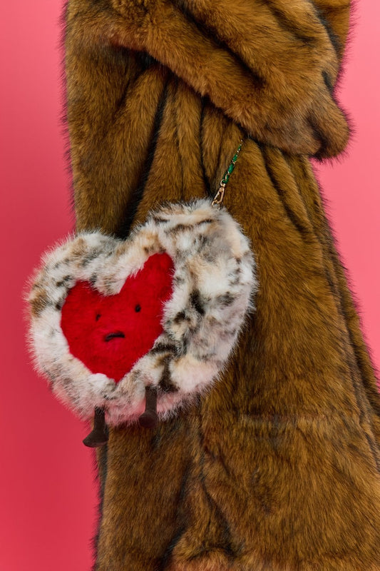 A person in a brown faux fur coat holds a faux fur heart bag with a red centre, small legs, and a face, standing against a pink background.