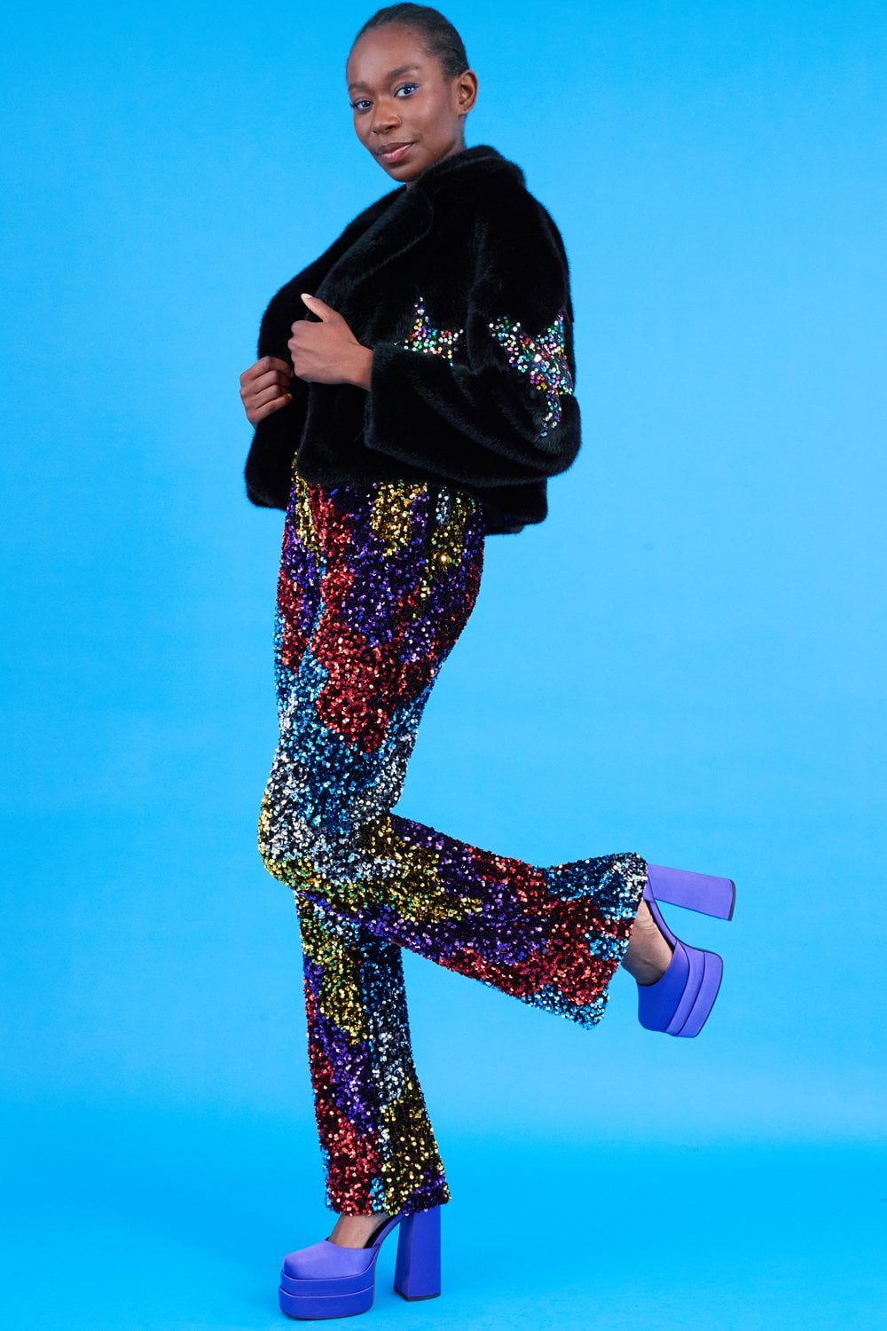 Person standing on one leg before a blue background, dressed in a black bamboo jacket with faux fur, multicoloured sequin star trousers, and purple platform heels.