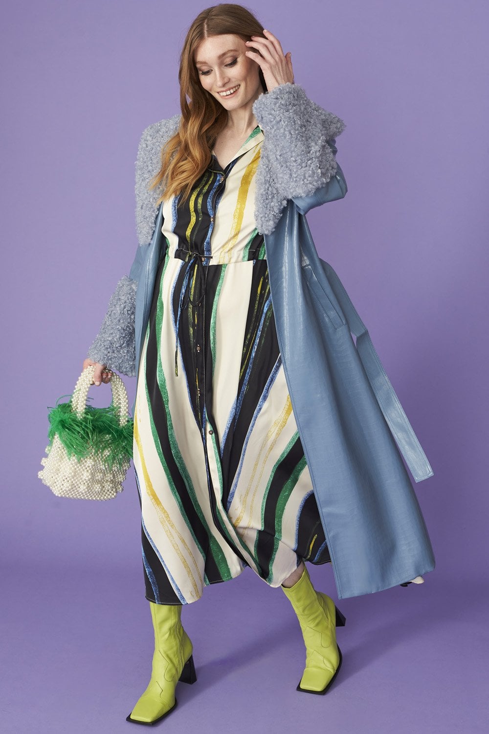 A woman stands against a purple background wearing a blue faux suede coat with detachable faux shearling cuffs, a striped dress, neon yellow boots, and holding a pearl handbag adorned with green feathers.