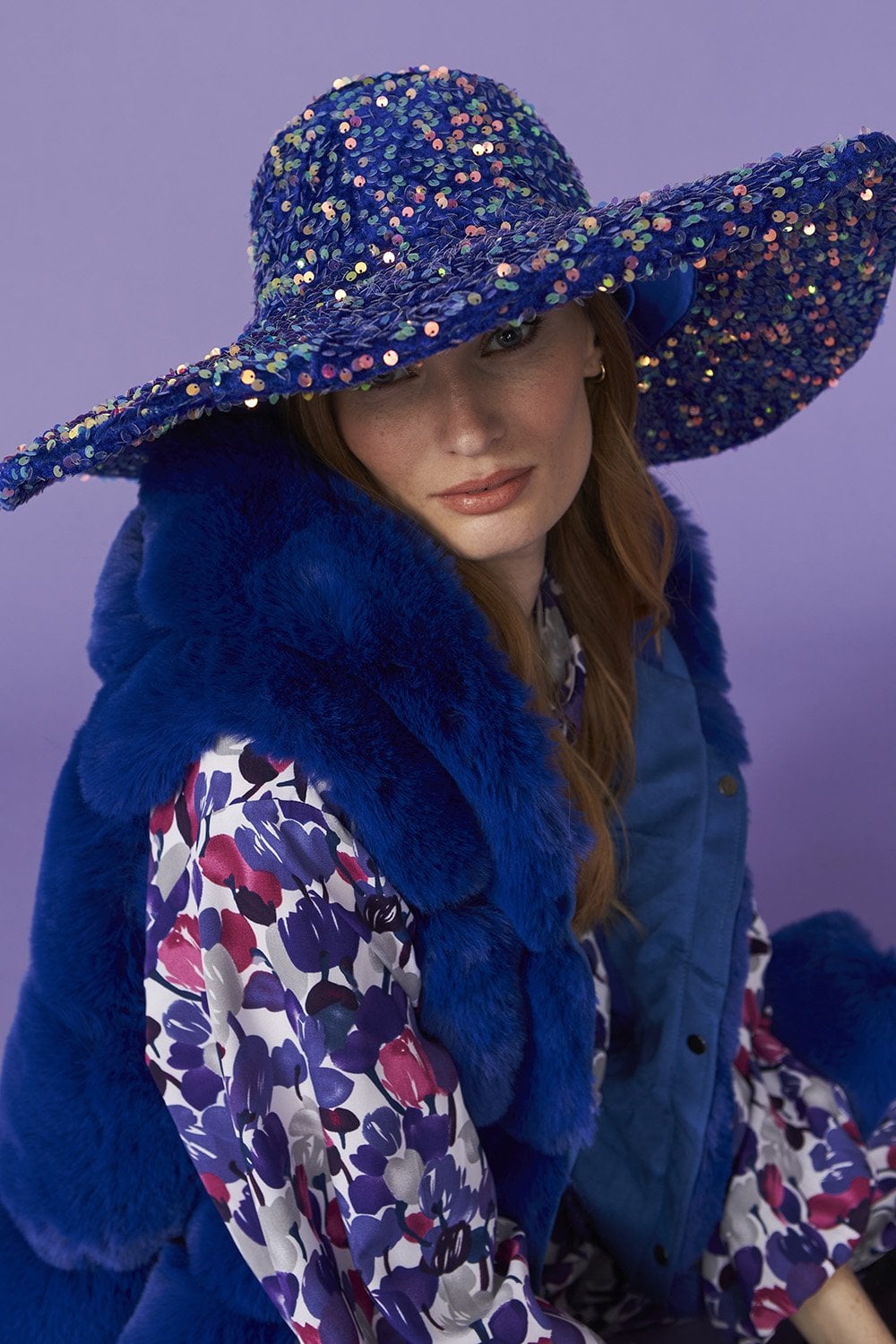 Woman wearing a wide-brimmed, blue sequin flapper hat, a blue fur gilet, and a floral patterned blouse, seated in front of a purple background for a touch of vintage glamour.