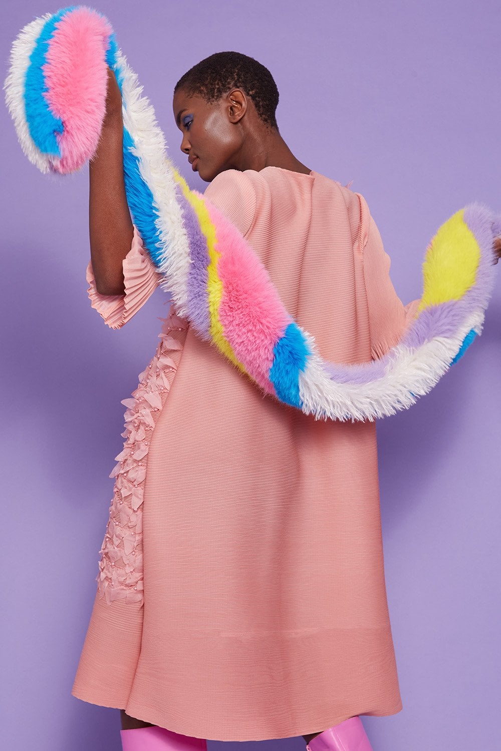 A person in a pale pink dress with frill details holds a long, sustainable faux fur scarf against a purple background.