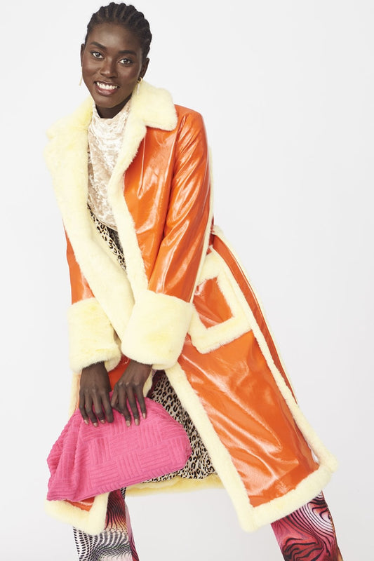 A person wearing an orange faux suede trench coat with yellow faux fur trim, patterned trousers, and holding a pink clutch smiles at the camera in front of a plain white background.