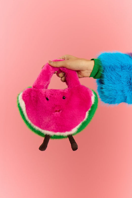A person wearing a colourful faux fur sleeve holds a plush watermelon bamboo bag shaped like a pink slice with a face and legs, set against a pink background.
