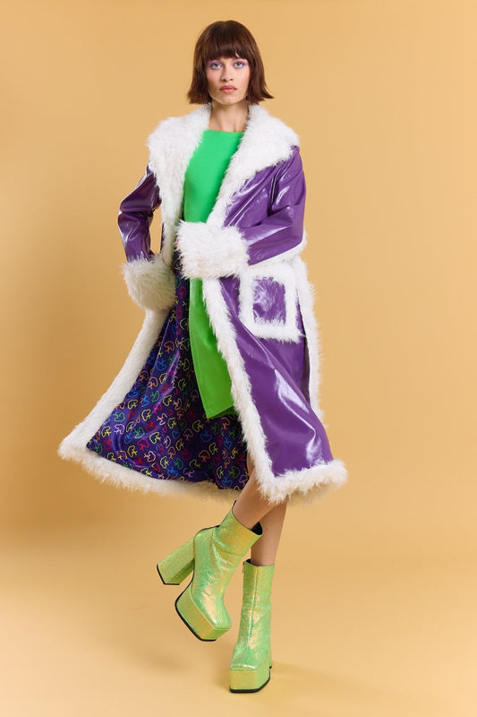 A person in a green dress, faux leather maxi coat with purple faux fur trim, and shiny green platform boots poses against a plain beige background.