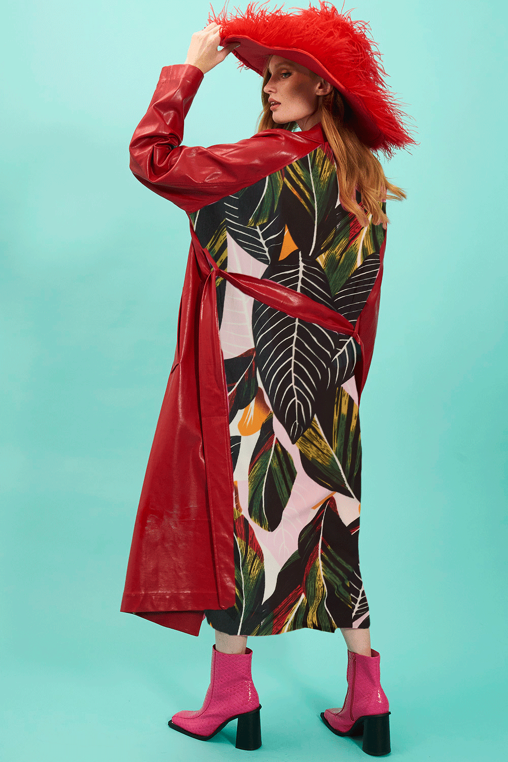 A woman stands against a turquoise background, wearing a red eco leather trench coat adorned with feathers, a tropical print dress, and pink boots.