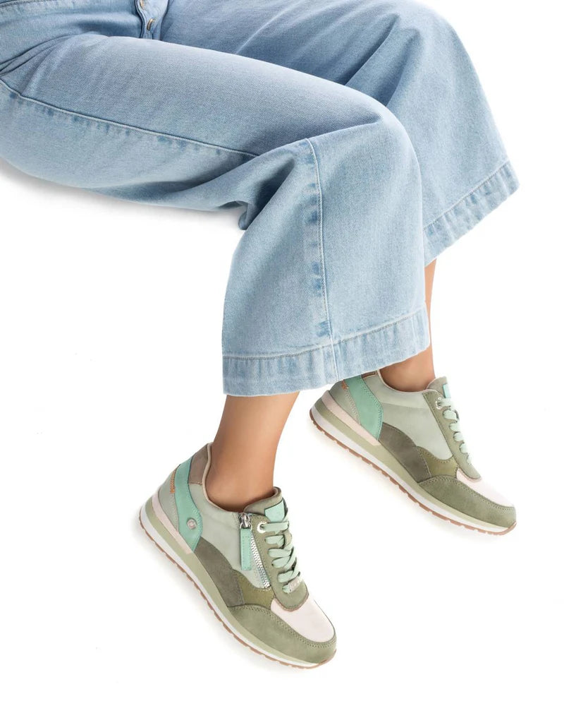A person wearing light blue wide-leg jeans and green vegan-friendly trainers is seated with their legs crossed at the ankles against a white background.