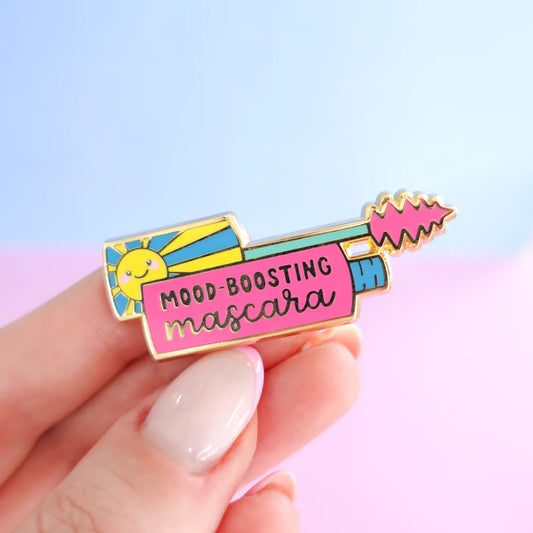 A hand holds a Mascara pin shaped like a mascara tube, featuring the words Mood-Boosting Mascara and a sun illustration—a delightful self-care gift set against a pastel-coloured background.
