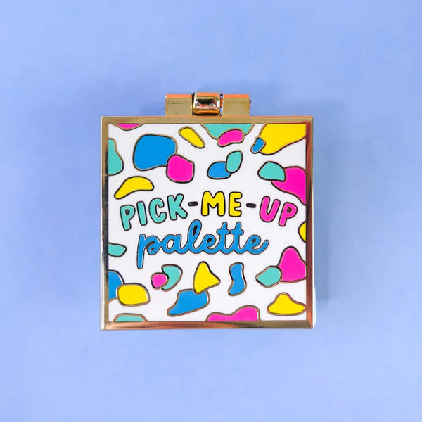 A square make-up palette with a gold border and colourful abstract shapes on the lid, labelled PICK-ME-UP palette, ideal as a self-care gift, set against a light blue background.