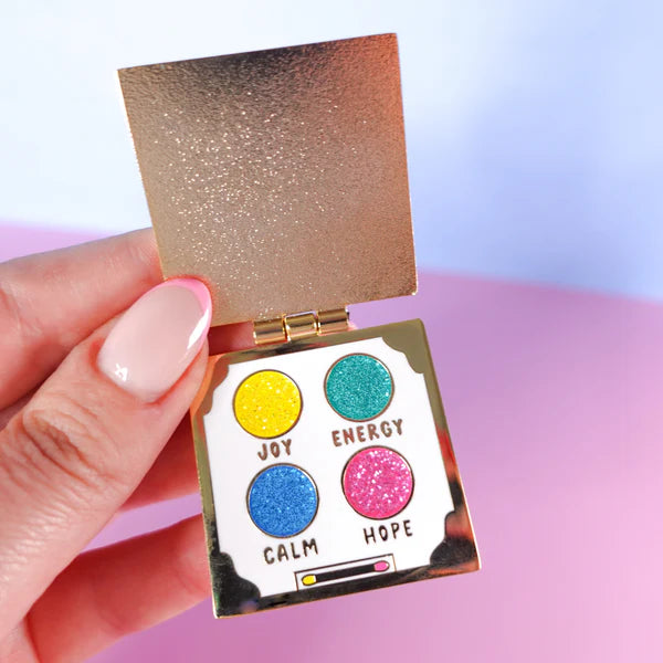 A hand holds an open, gold-coloured eyeshadow palette pin featuring four glittery circles labelled Joy (yellow), Energy (teal), Calm (blue), and Hope (pink)—a delightful self-care gift.