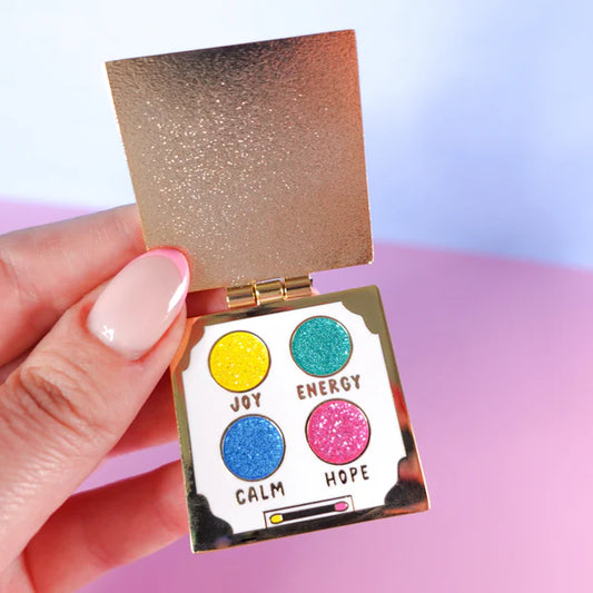 A hand holds an open, gold-coloured eyeshadow palette pin featuring four glittery circles labelled Joy (yellow), Energy (teal), Calm (blue), and Hope (pink)—a delightful self-care gift.