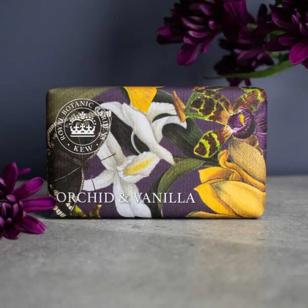 A wrapped Vegan Orchid and Vanilla Soap Bar adorned with floral illustrations rests on a grey surface next to purple flowers, emphasising its gentle and eco-friendly qualities.
