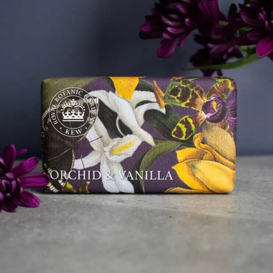 A wrapped Vegan Orchid and Vanilla Soap Bar adorned with floral illustrations rests on a grey surface next to purple flowers, emphasising its gentle and eco-friendly qualities.