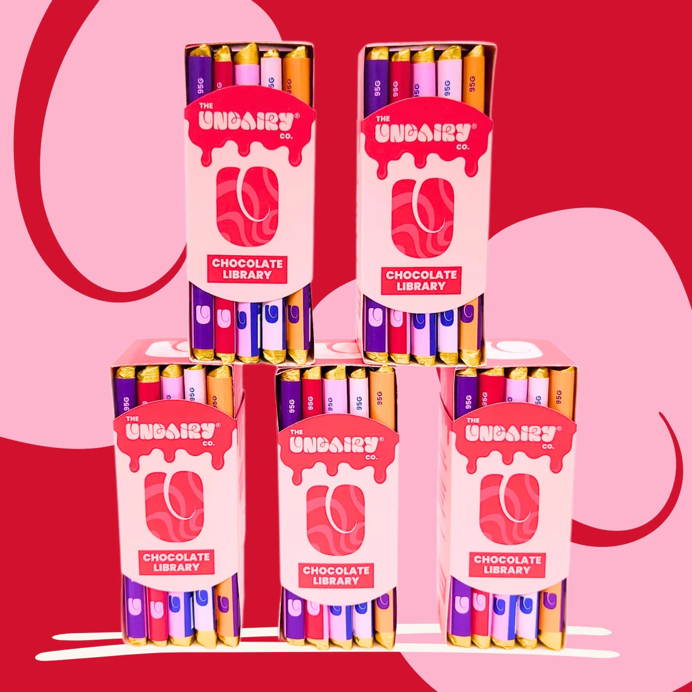 Five boxes of The UnDairy Co. Chocolate Library, featuring the Original Creamy bar and Fruit & Nut bar, are stacked in a pyramid against a striking red and pink patterned background.