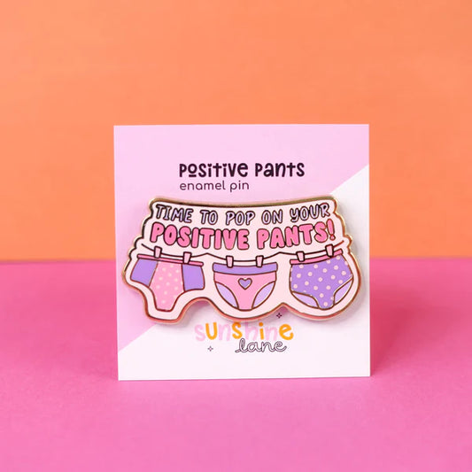 An enamel pin depicting illustrated pants hanging on a washing line, with the text Time to pop on your positive pants! A cheerful and uplifting gift set against a lively pink and orange background.
