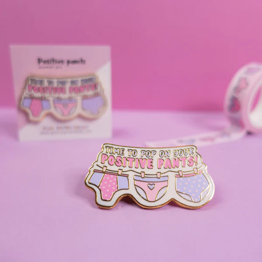 A colourful enamel pin featuring illustrations of pants on a washing line and the text "Time to pop on your positive pants!"—an uplifting gift shown against a pink background.