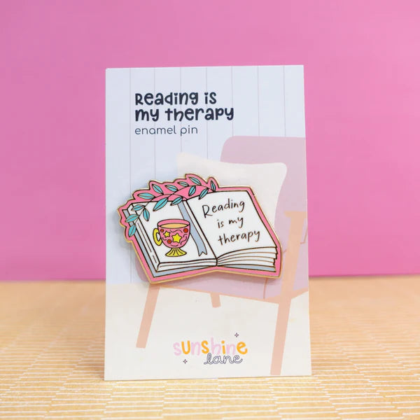 Enamel pin in the shape of an open book with a cup and flowers, bearing the words Reading is my therapy, displayed on a card against a pink background—a charming reading accessory and ideal gift for book lovers.