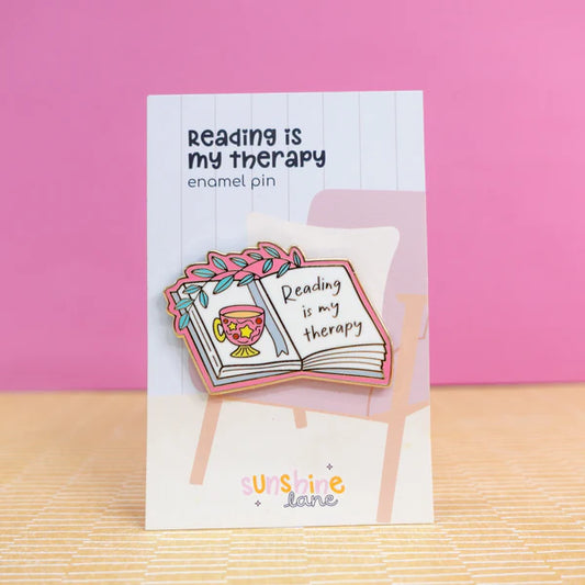 Enamel pin in the shape of an open book with a cup and flowers, bearing the words Reading is my therapy, displayed on a card against a pink background—a charming reading accessory and ideal gift for book lovers.