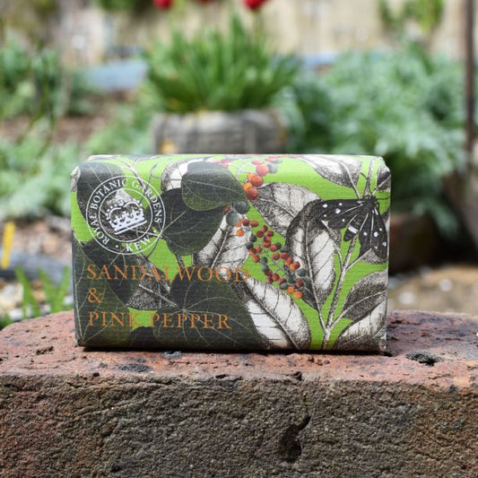 A bar of vegan sandalwood soap wrapped in green and botanical-print paper labelled Sandalwood & Pink Pepper rests on a stone outdoors, with plants in the background.