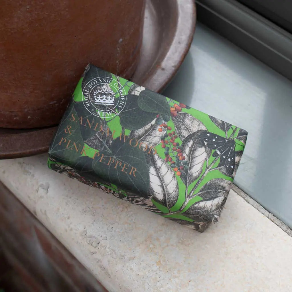 A vegan sandalwood soap, wrapped in green and black botanical paper labelled “Sandalwood & Pink Pepper”, sits beside a brown pot on the windowsill.