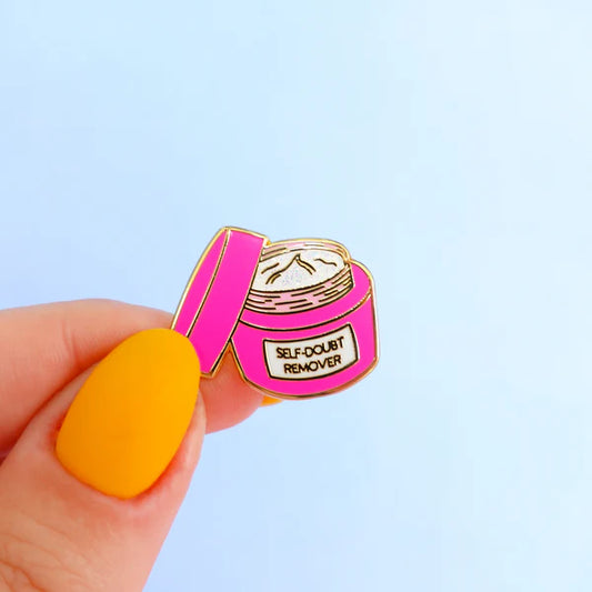 A hand with yellow nail varnish holds a self-doubt remover pin—an enamel pin in the shape of a pink jar labelled SELF-DOUBT REMOVER—set against a pale blue background.