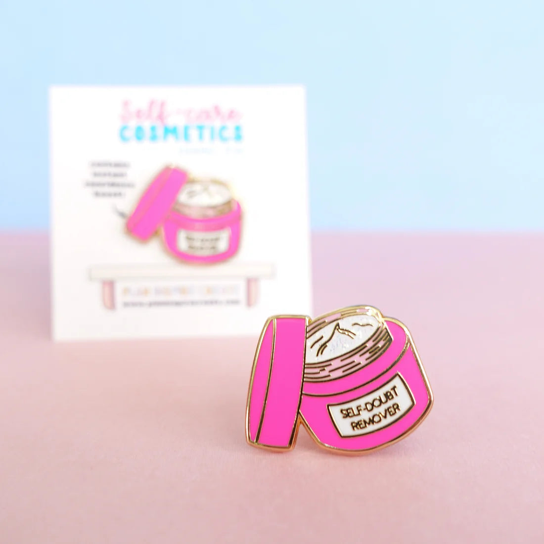 A pink enamel pin shaped like an open cosmetics jar, labelled SELF-DOUBT REMOVER, rests on a pink surface alongside a matching card—a perfect confidence-boosting pin for your collection.