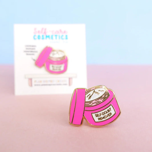 A pink enamel pin shaped like an open cosmetics jar, labelled SELF-DOUBT REMOVER, rests on a pink surface alongside a matching card—a perfect confidence-boosting pin for your collection.
