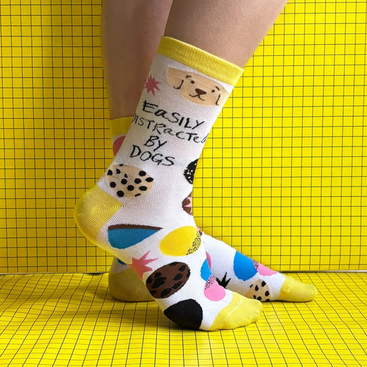 Individual wearing colourful dog lover socks featuring dog illustrations and the phrase "Easily distracted by dogs" set against a yellow, grid-patterned background.