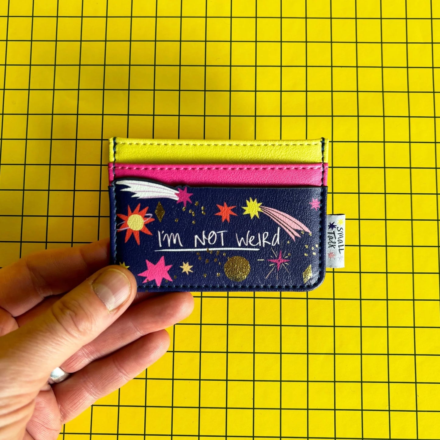 A hand holds a quirky cardholder decorated with stars and the phrase IM NOT WEIRD, against a yellow grid background.