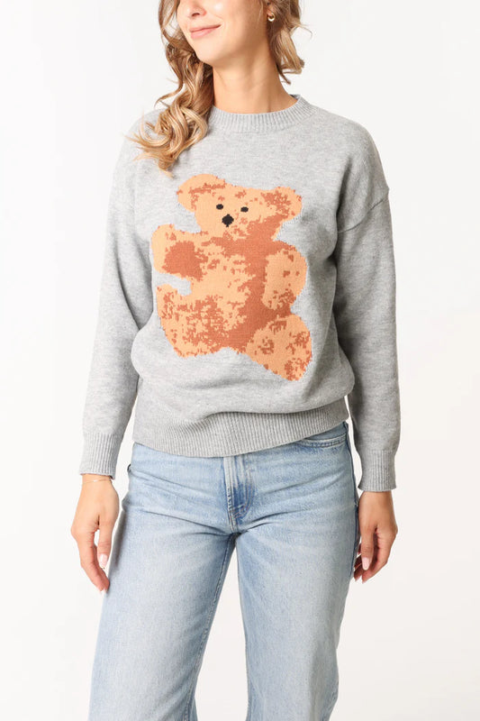 A person in a cosy bear jacquard jumper featuring a large teddy graphic on the front, paired with light blue jeans, stands against a plain background.