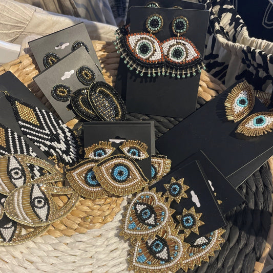 Several pairs of handcrafted earrings, including Mini Gold Beaded Eye Stud Earrings with geometric patterns and surgical steel posts, are displayed on black cards in a woven basket.