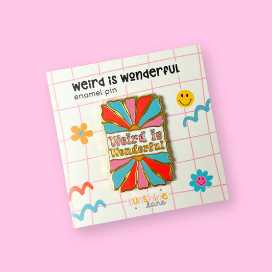 A colourful gold enamel pin on a pink grid card reads ‘Weird is Wonderful’, featuring abstract boho flower shapes and small doodles of a smiley face, heart, and clouds.
