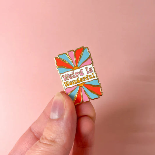 A hand holds a colourful gold enamel pin bearing the words "Weird is Wonderful" against a light pink background.