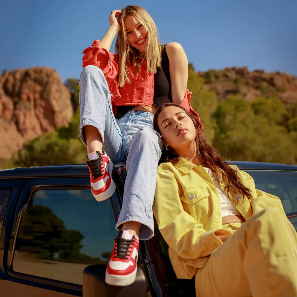 Two women sit on the bonnet of a car in a sunny outdoor setting; one wears a red top and jeans, while the other wears a yellow jacket and trousers, sporting stylish vegan trainers. A rocky landscape and trees are in the background.