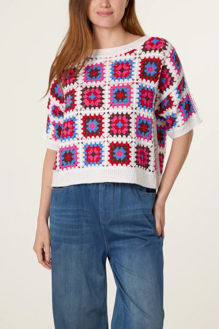 A woman is wearing a short-sleeved, multicoloured patchwork crochet top with white borders and blue jeans, standing against a plain background.
