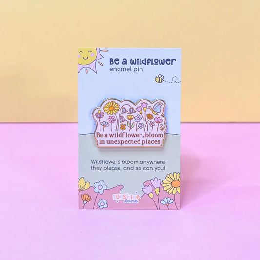 A colourful floral enamel pin featuring flowers and the phrase 'Be a wildflower, bloom in unexpected places', presented on a pastel card adorned with delightful flower and sun illustrations.