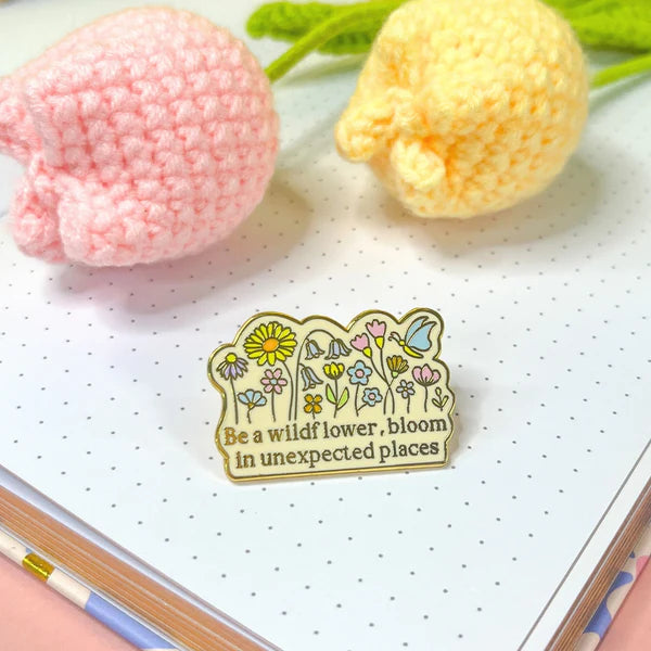 A wildflower badge bearing the phrase Be a wildflower, bloom in unexpected places rests on a dotted notebook next to two crocheted flowers.