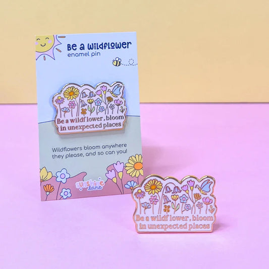 A floral enamel pin featuring the phrase Be a wildflower, bloom in unexpected places is shown before a matching decorative card on a pastel pink and purple background—the ideal wildflower pin for any collection.