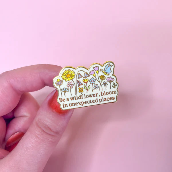 A hand holds a wildflower pin with illustrated blooms and the words, Be a wildflower, bloom in unexpected places, set against a pink background.