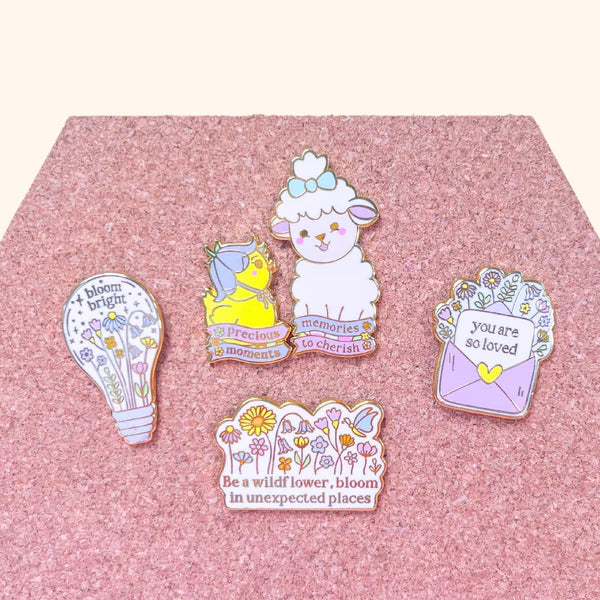 Five colourful enamel pins, including a floral pin and a wildflower pin with positive messages, are arranged on a pink corkboard. The pins depict flowers, a sheep, a bird, a lightbulb, and uplifting text.