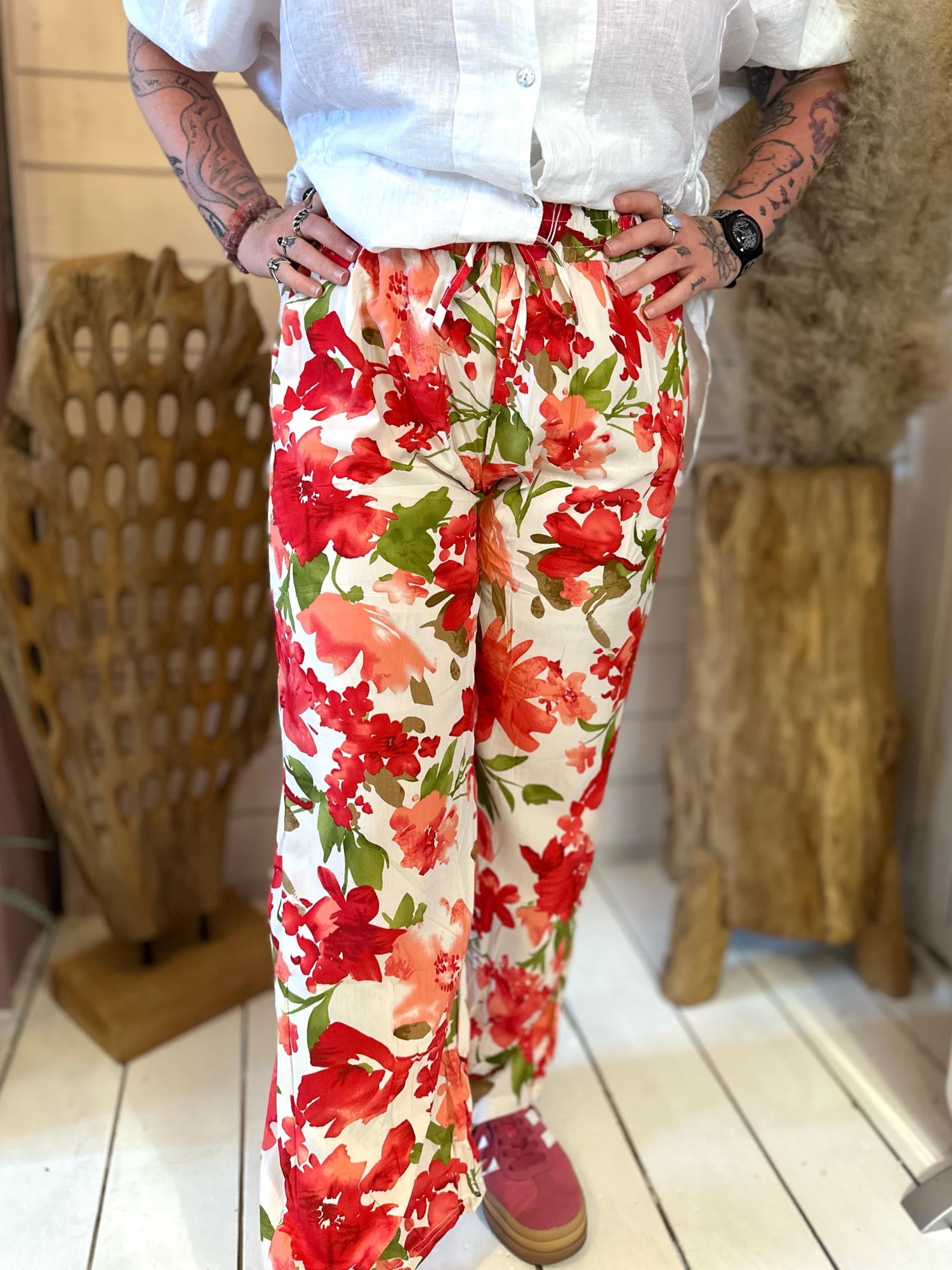 Person standing indoors wearing a white shirt and loose, high-waisted trousers with a red and green floral pattern; hands on hips, face not visible.