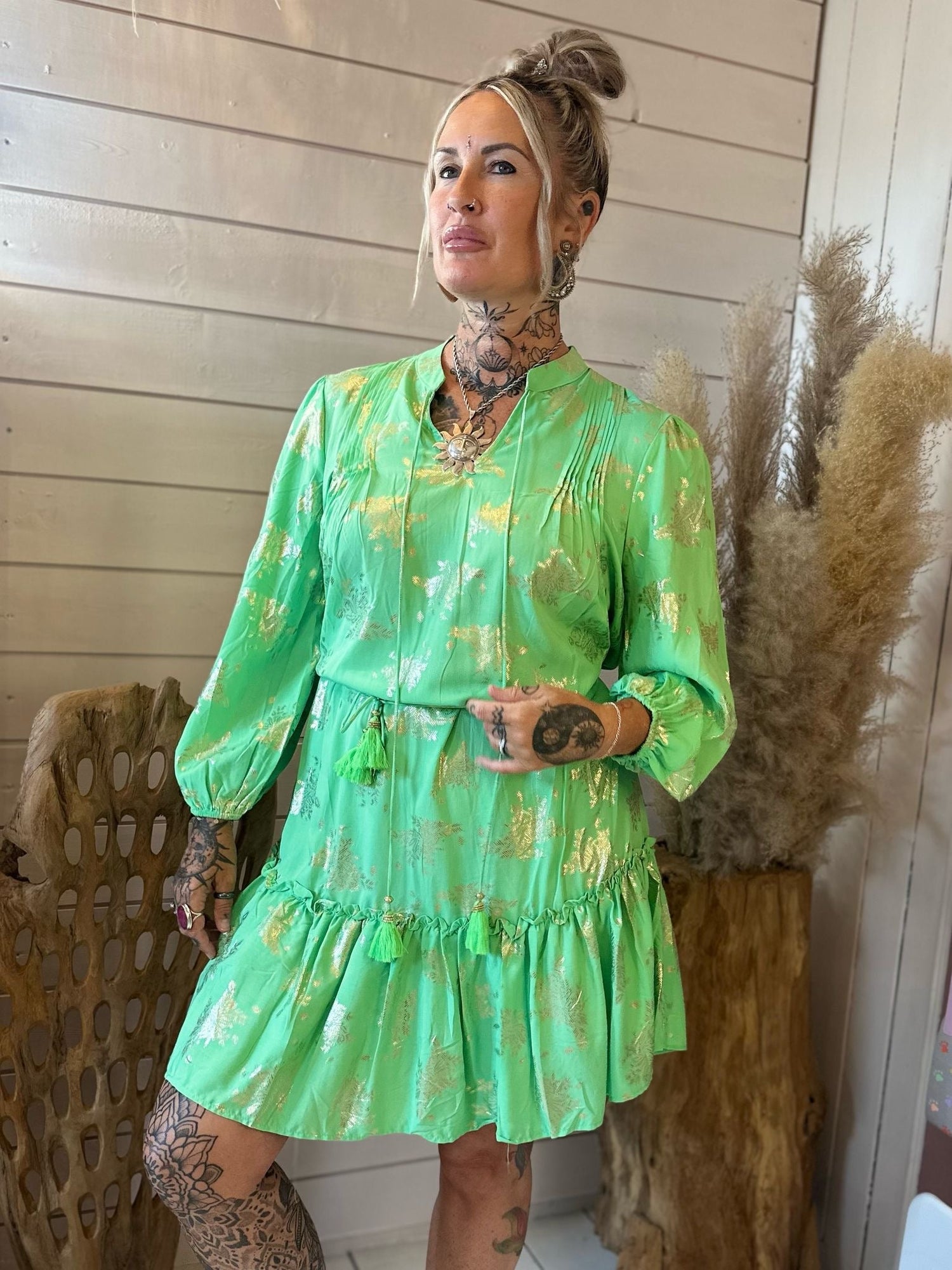 A woman with tattoos poses indoors, wearing a shiny green dress with gold patterns. She stands in front of a white wooden wall and dried pampas grass.