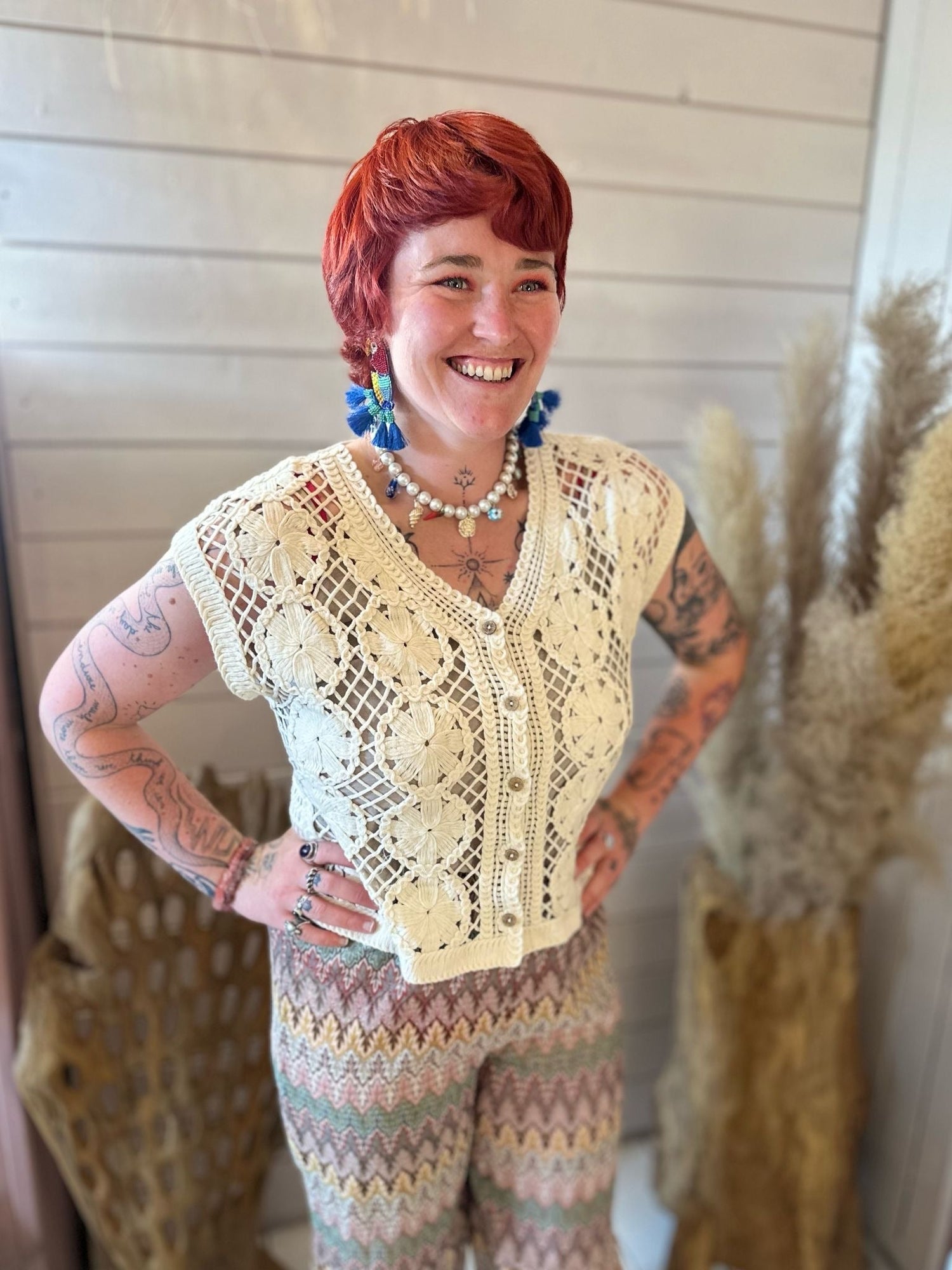 A person with short red hair smiles while wearing a cream crochet top and patterned trousers, standing indoors beside decorative dried plants.