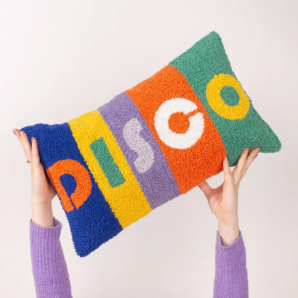 A person in a purple jumper holds a rectangular cushion with textured, colourful stripes spelling DISCO in bold letters.