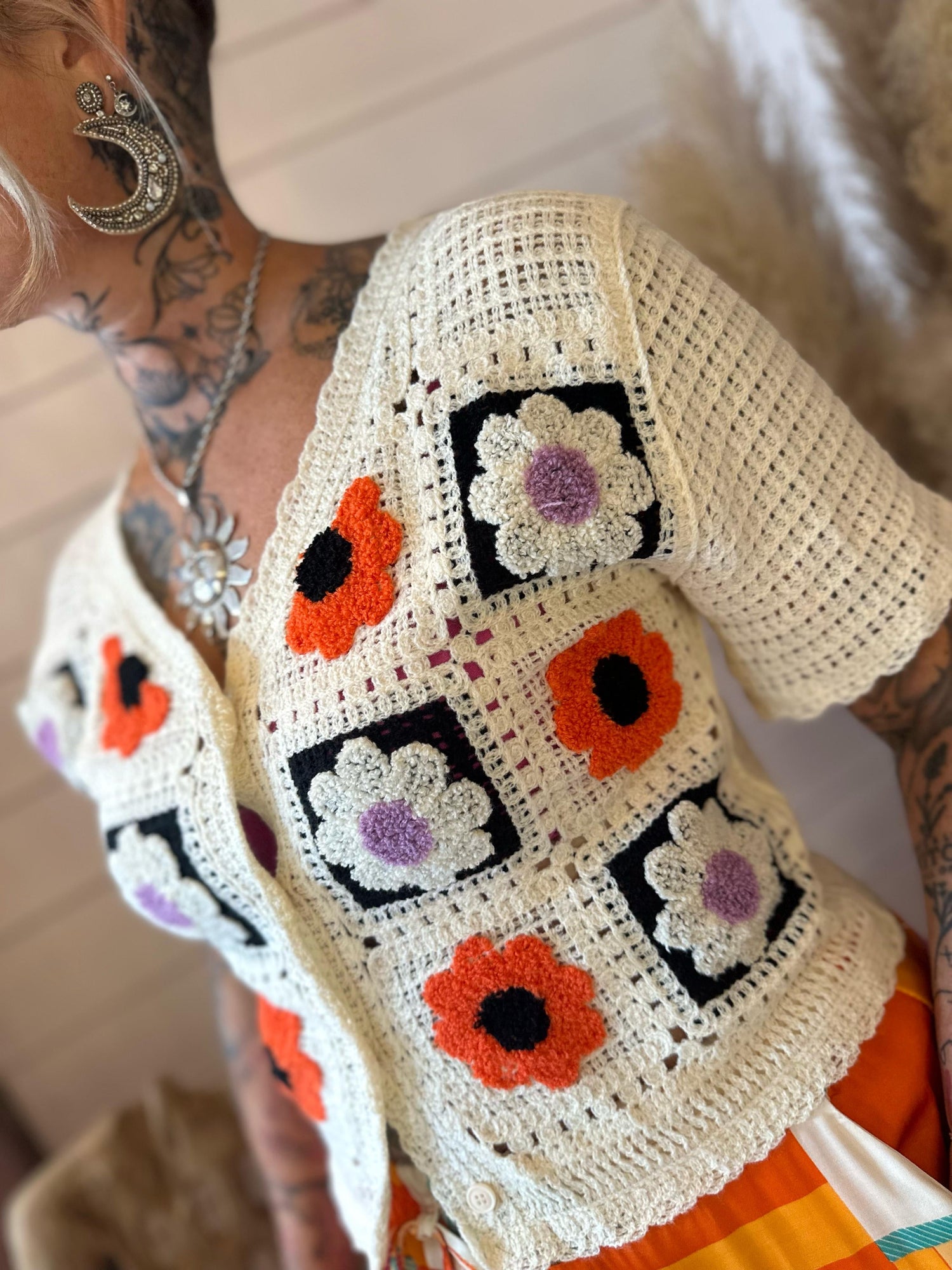 A person wearing a cream-coloured crocheted cardigan with orange and purple floral square patterns, featuring intricate tattoos and jewellery.
