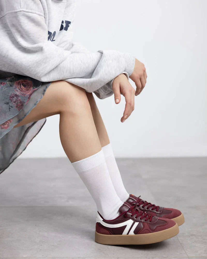 Person wearing a grey sweatshirt, floral skirt, white knee-high socks, and burgundy trainers sits on a grey floor with legs bent and arms resting on knees.