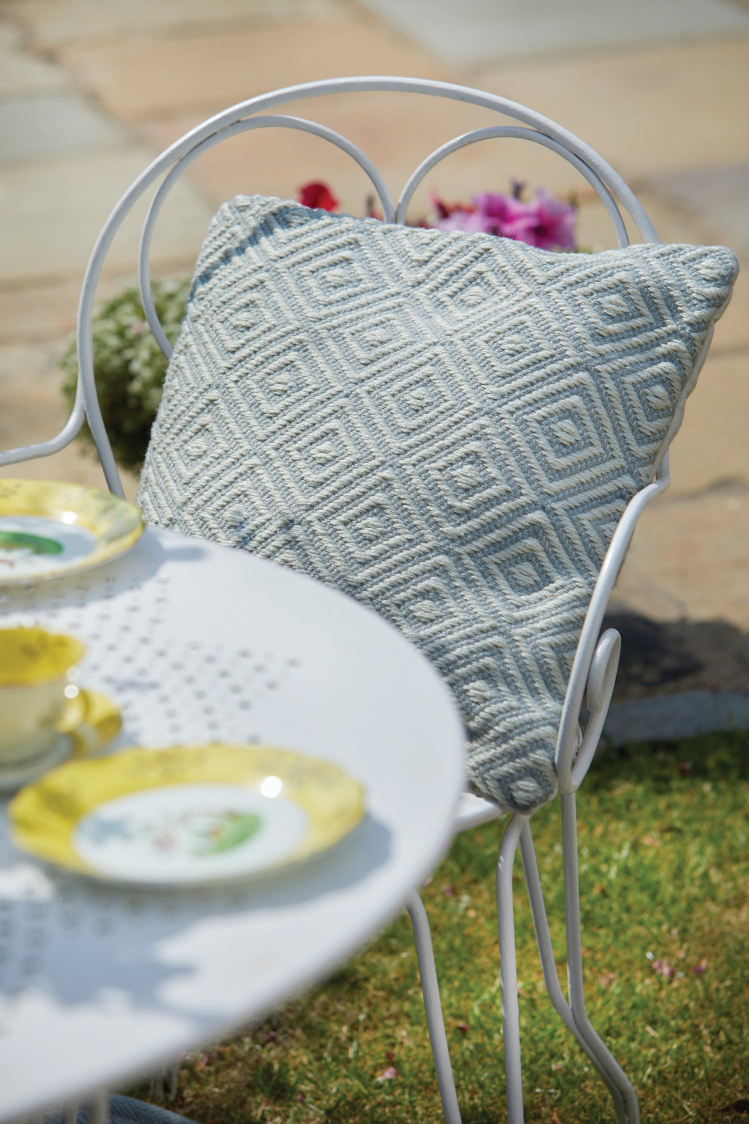 A white metal chair with a grey, textured geometric cushion sits next to a round table laid with pastel teacups and plates in a garden setting.