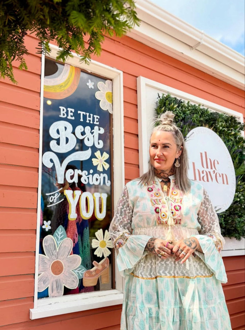 A woman wearing a Jessie short dress with three-quarter sleeves stands in front of a peach-coloured building with a sign reading “Be the Best Version of You” and another sign that says “The Haven.”.